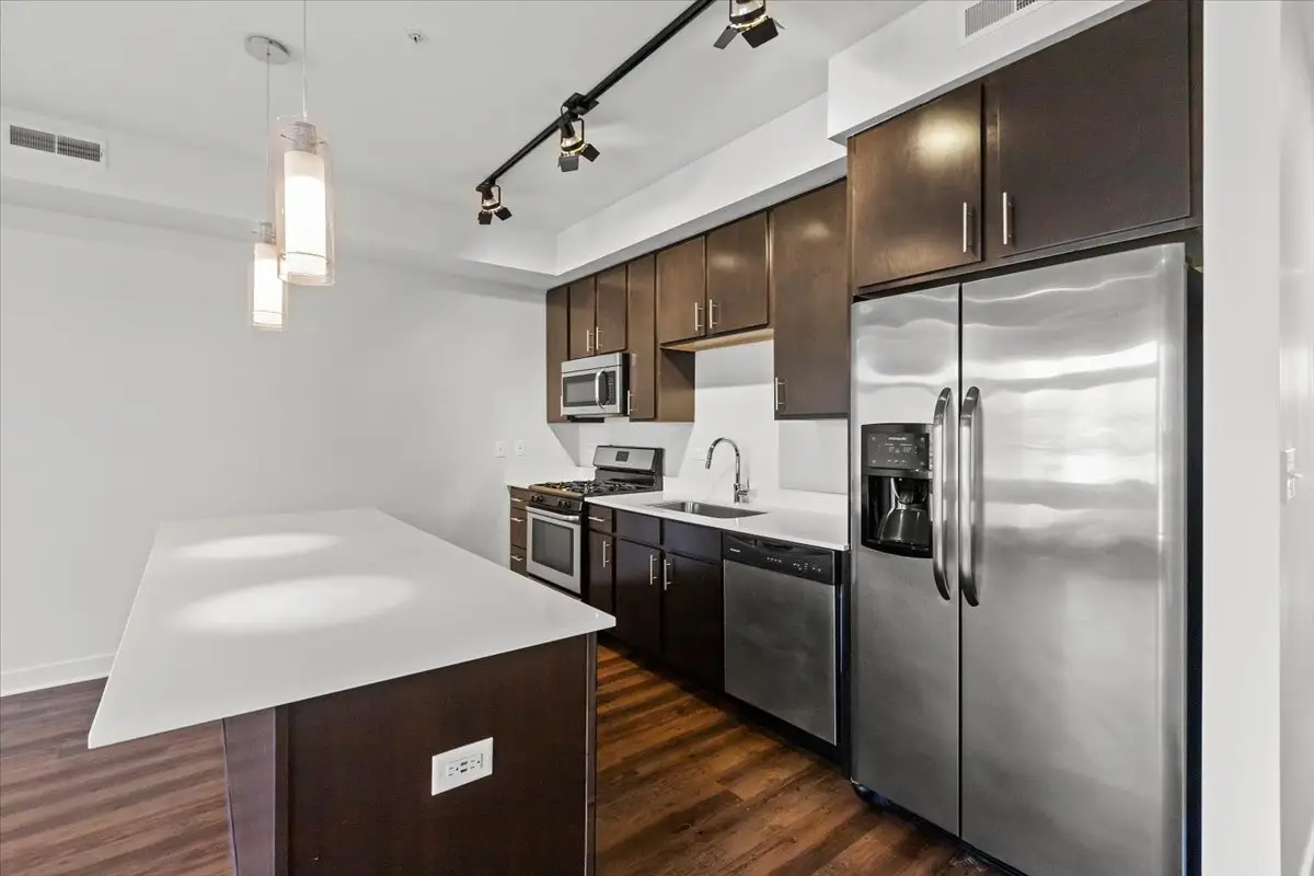 Open layout kitchen with pendant lights over a large island, white hard stone countertops, large stainless refrigerator, espresso cabinets with brushed nickel hardware, and overhead track lighting.