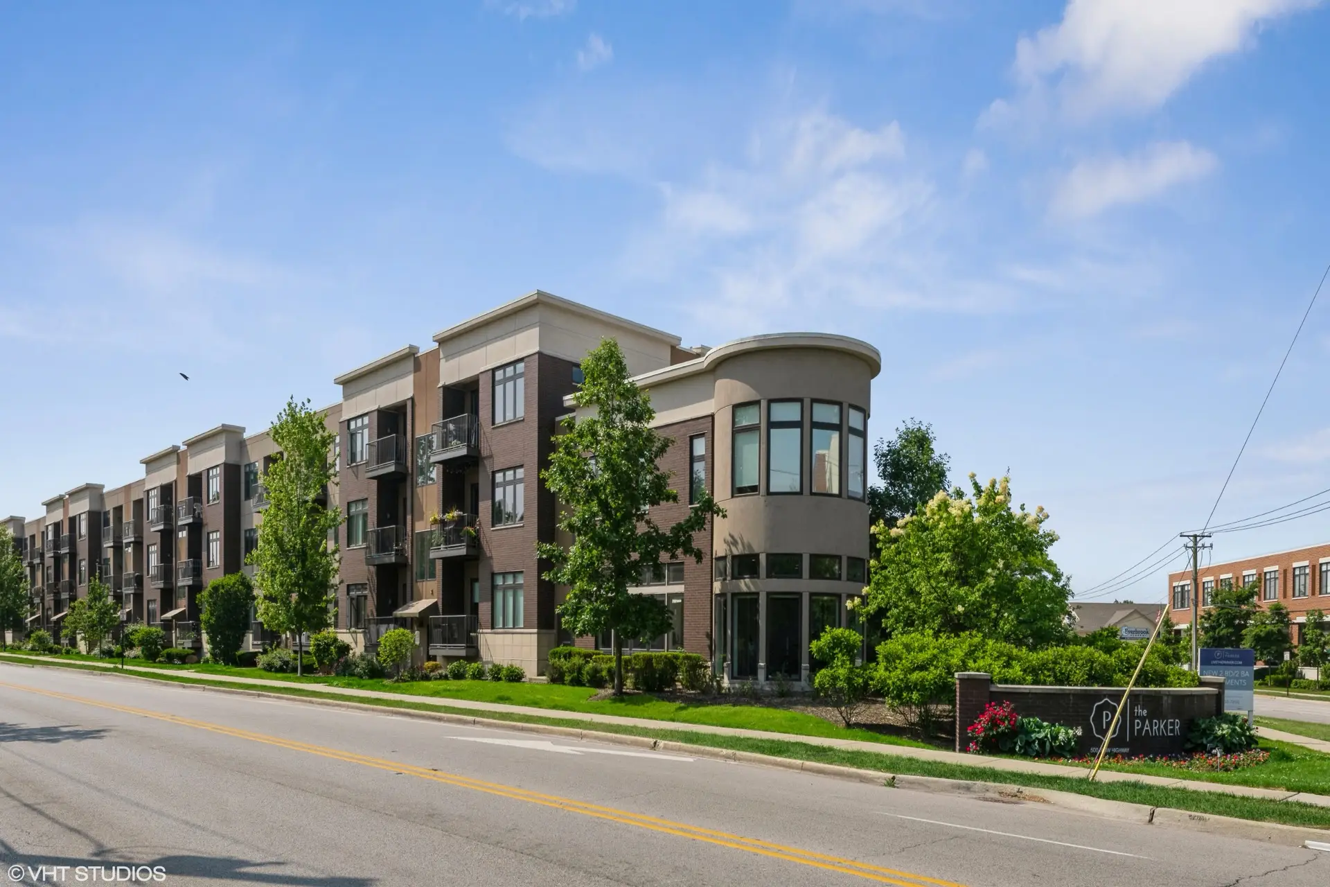 The Parker three story apartment building exterior of red brick and tan flat stone viewed at an angle from the street with several green trees along the side.