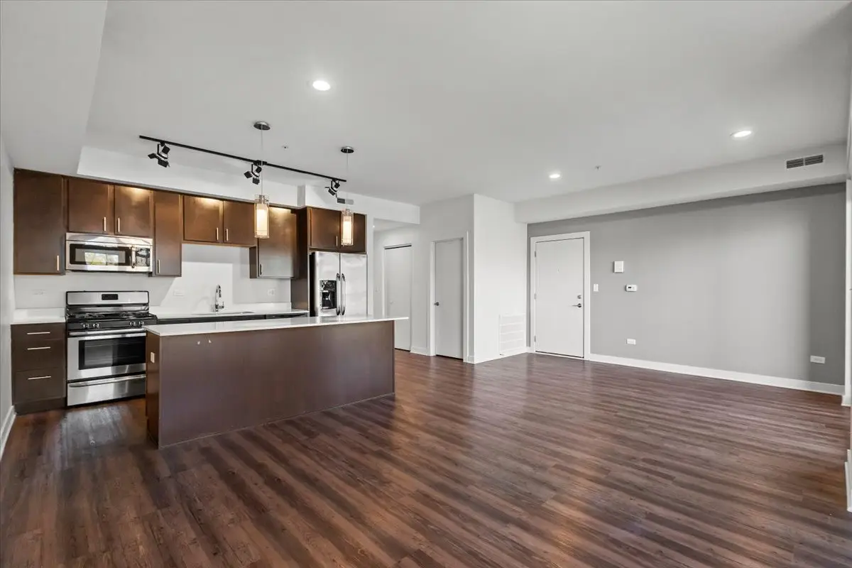 view from living room of open layout kitchen with large island, dark brown wood style flooring throughout, and can lights in the ceiling.