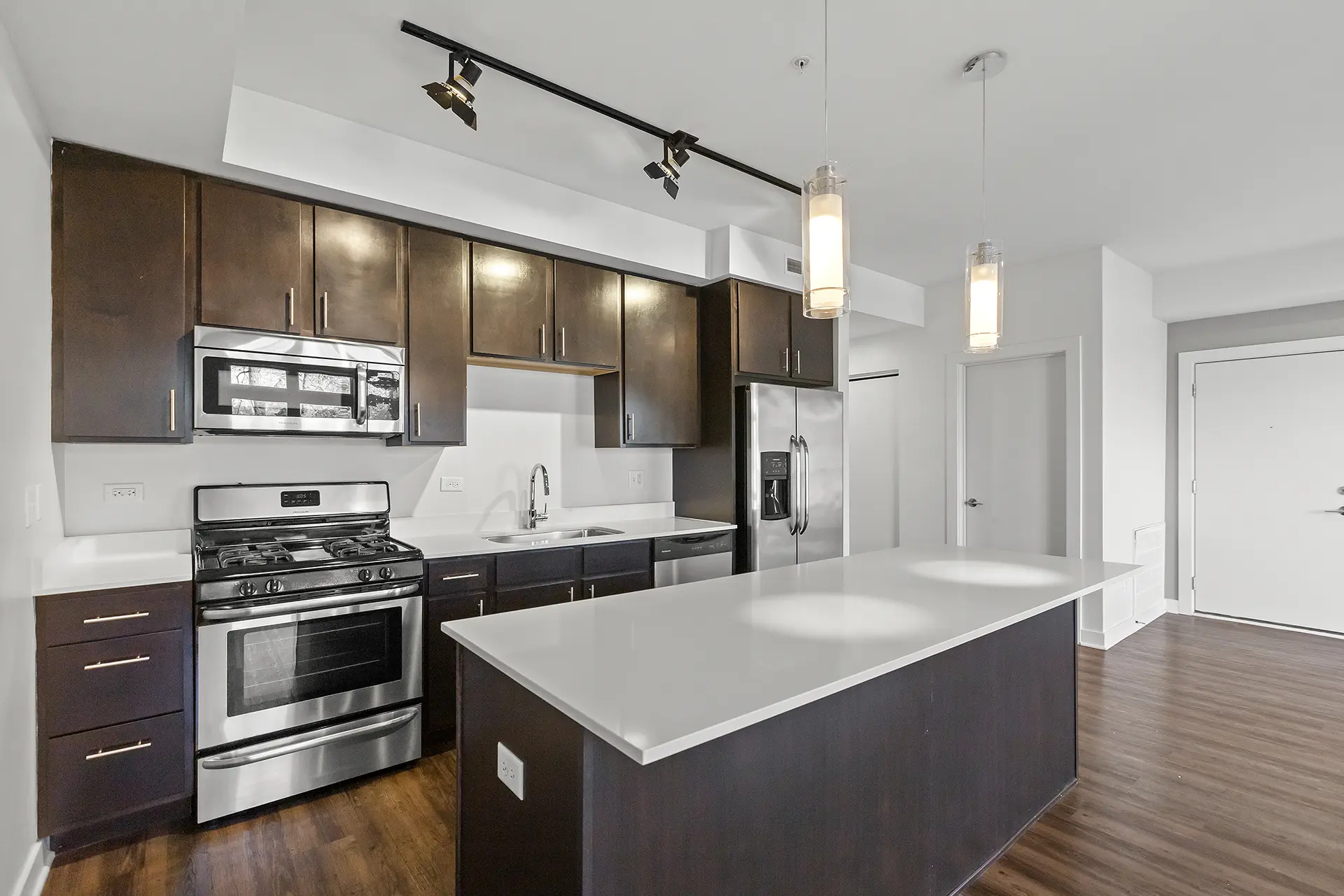 Open layout kitchen with pendant lights over a large island, white hard stone countertops, large stainless refrigerator, espresso cabinets with brushed nickel hardware, and overhead track lighting.