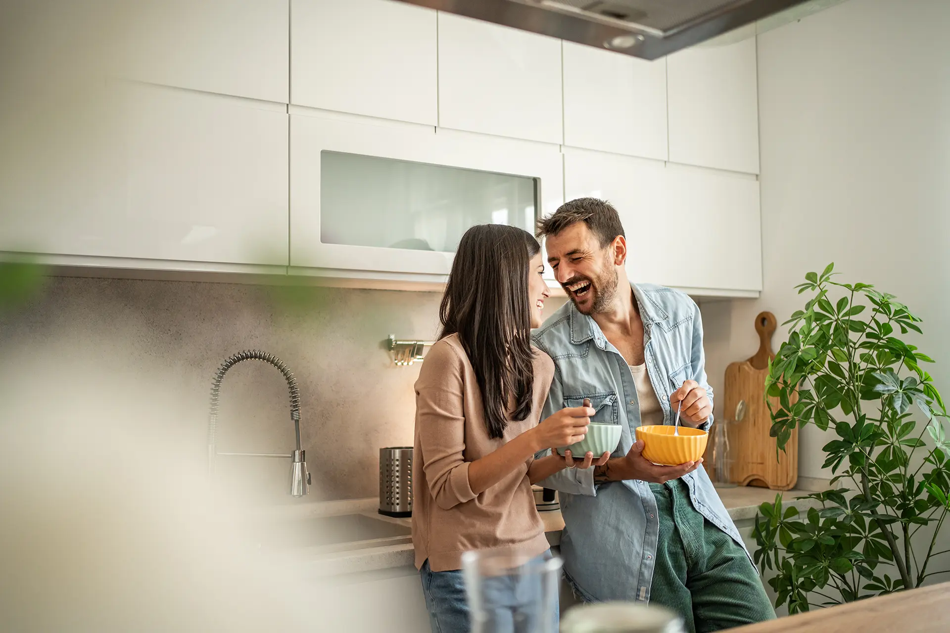 A young couple laughing and leaning against the kitchen sink countertop while holding blue and orange cereal bowls.