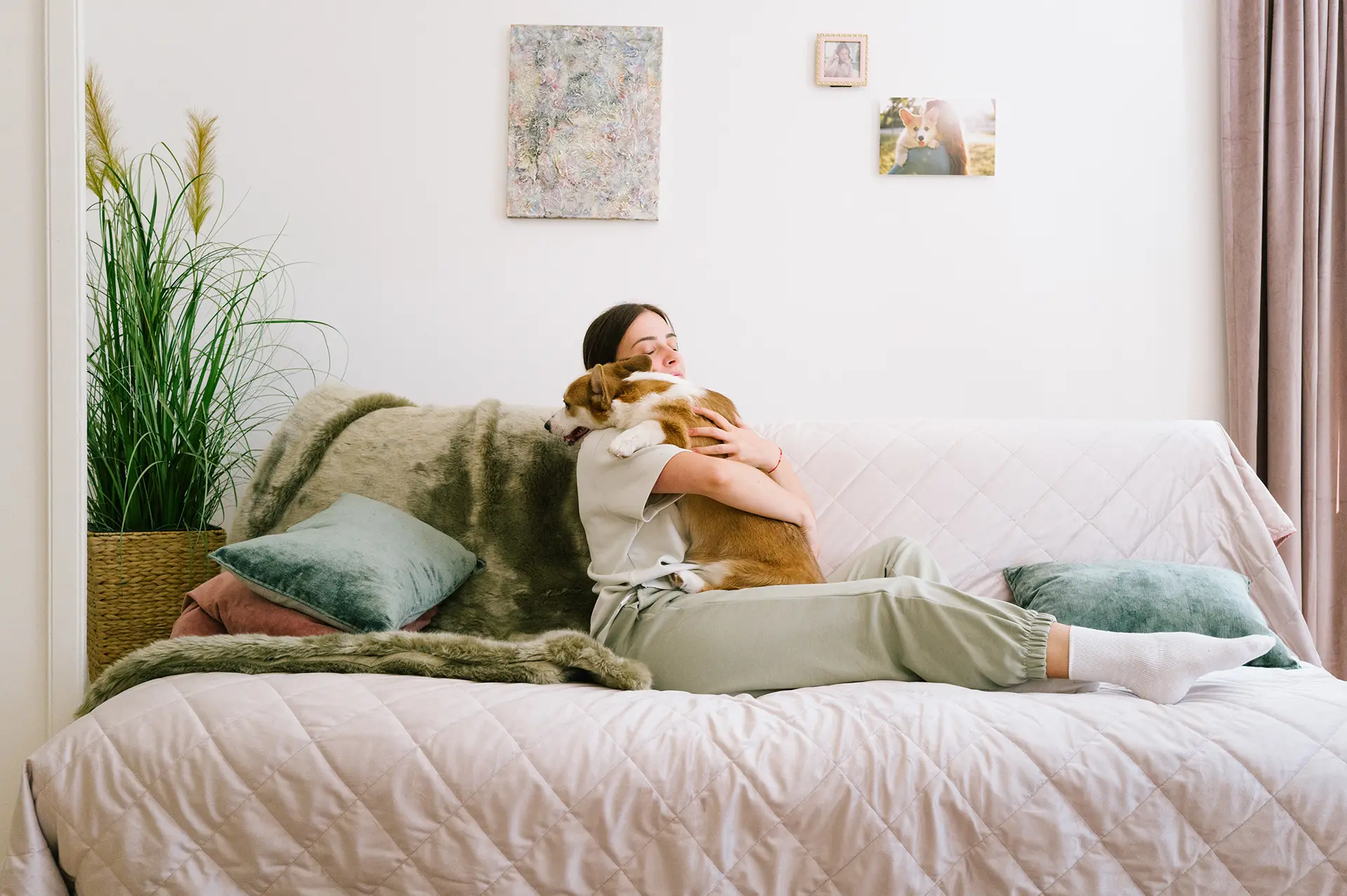 A young woman sitting up in bed hugging a corgi that is resting its head on her shoulder.